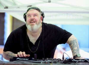 Kristian Nairn looking up wearing a black shirt and beaded necklace DJing.